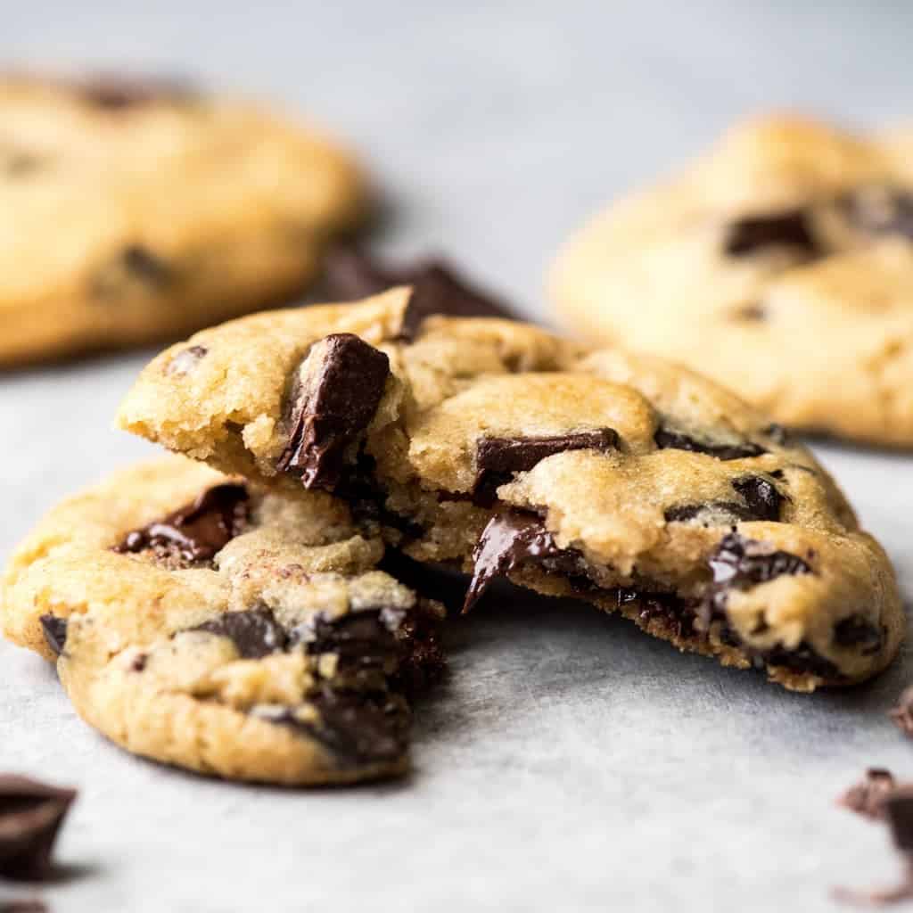 Front view of the best chocolate chip cookie recipe ever one cookie cut in half with melty chocolate chips surrounded by three other cookies and chopped chocolate