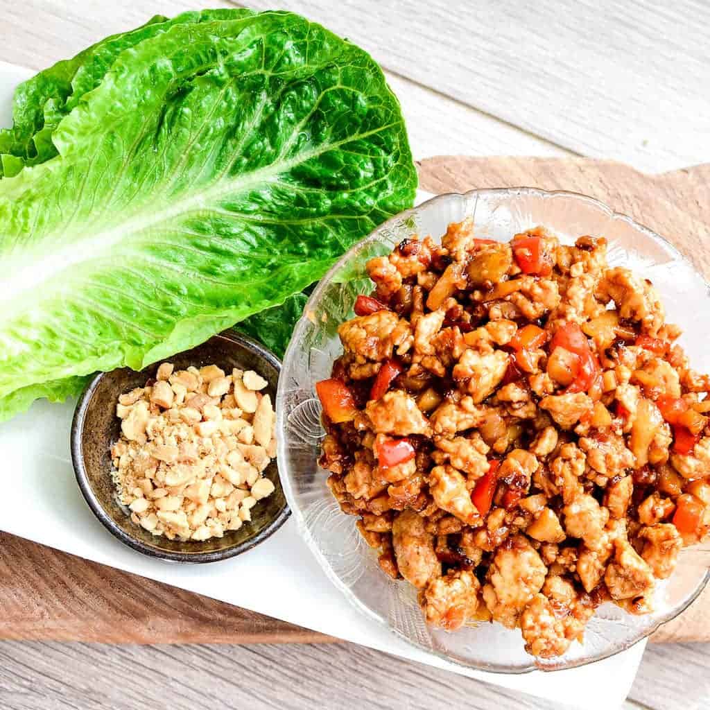 Overhead picture of asian chicken lettuce wrap filling in a bowl on a platter with fresh lettuce and crushed peanuts. 