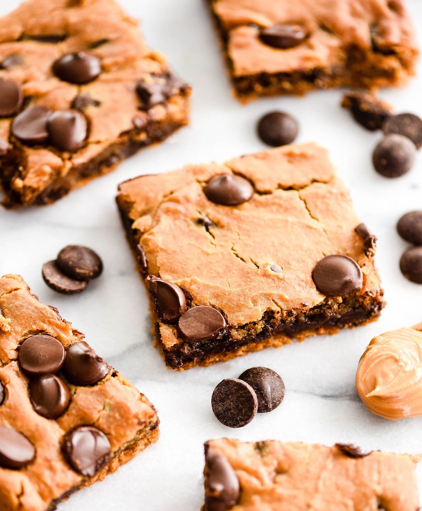 Angled overhead shot of one healthy peanut butter blondie surrounded by 4 other healthy peanut butter blondies, chocolate chips and a spoon full of peanut butter
