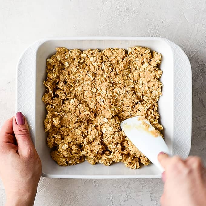 overhead view of a hand pressing the No-Bake Oatmeal Bars mixture into a white 8x8" baking dish with a white spatula 