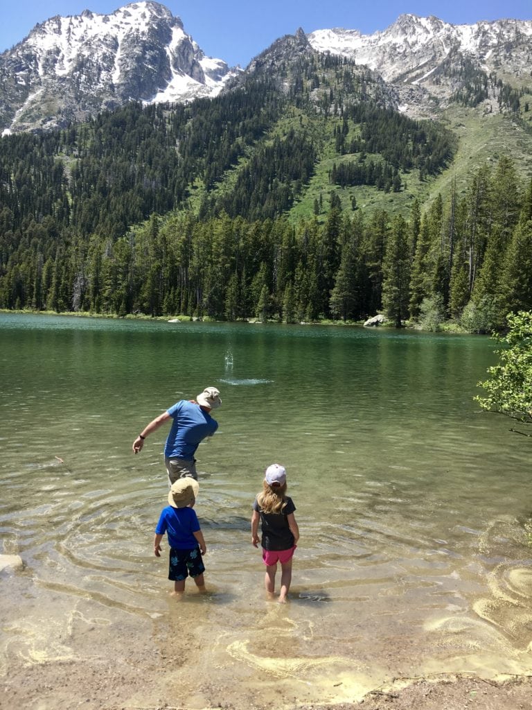 Family skipping rocks in a lake in the mountains in The Complete Guide to Hiking with Kids