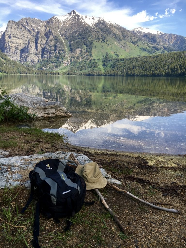 A photo of a backpack, hiking stick and hat laying near a lake in the mountains in The Complete Guide to Hiking with Kids