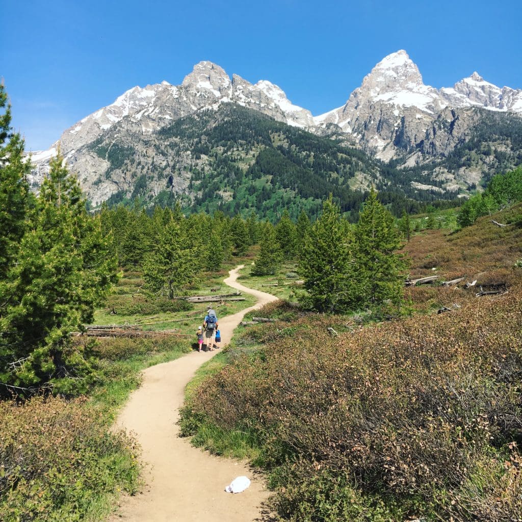 Photo of dad carrying a baby with two younger children on a hiking trail in the mountains in The Complete Guide to Hiking with Kids