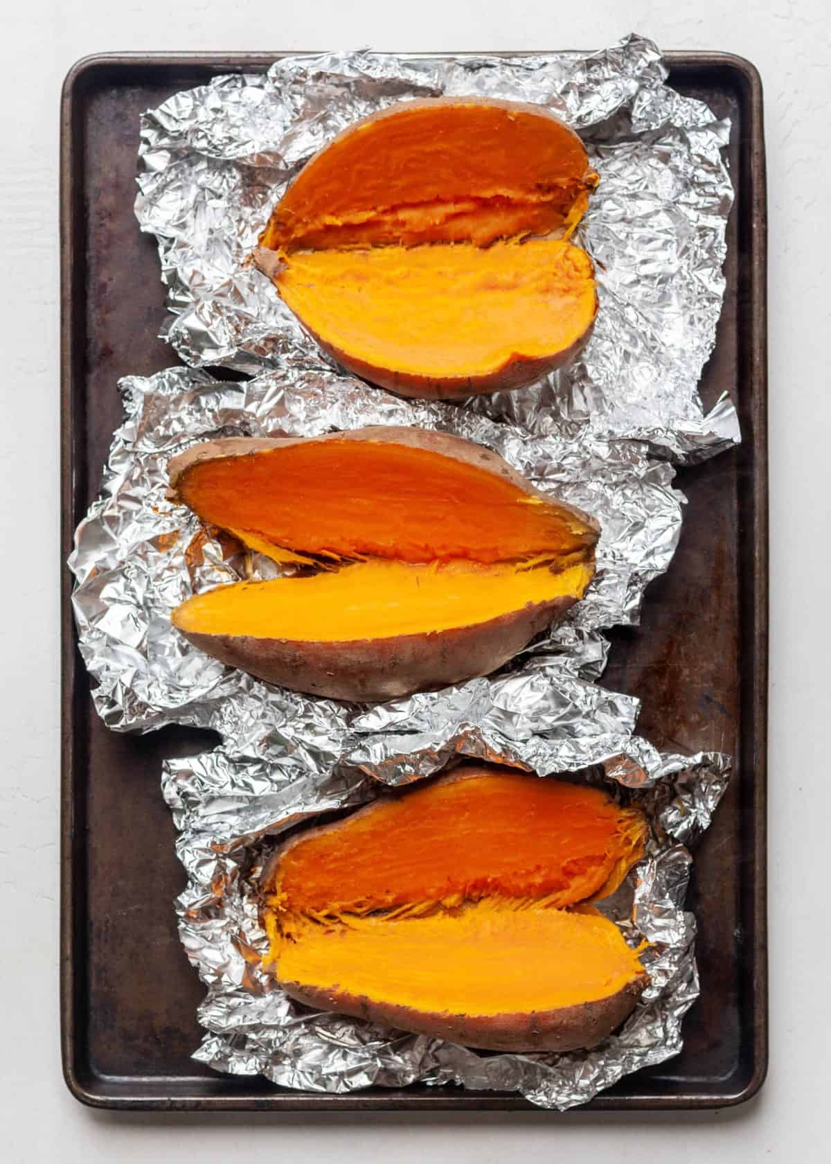 making healthy sweet potato casserole recipe - 3 baked sweet potatoes all sitting on foil on a baking sheet, all three are cut in half so the cooked flesh is visible.