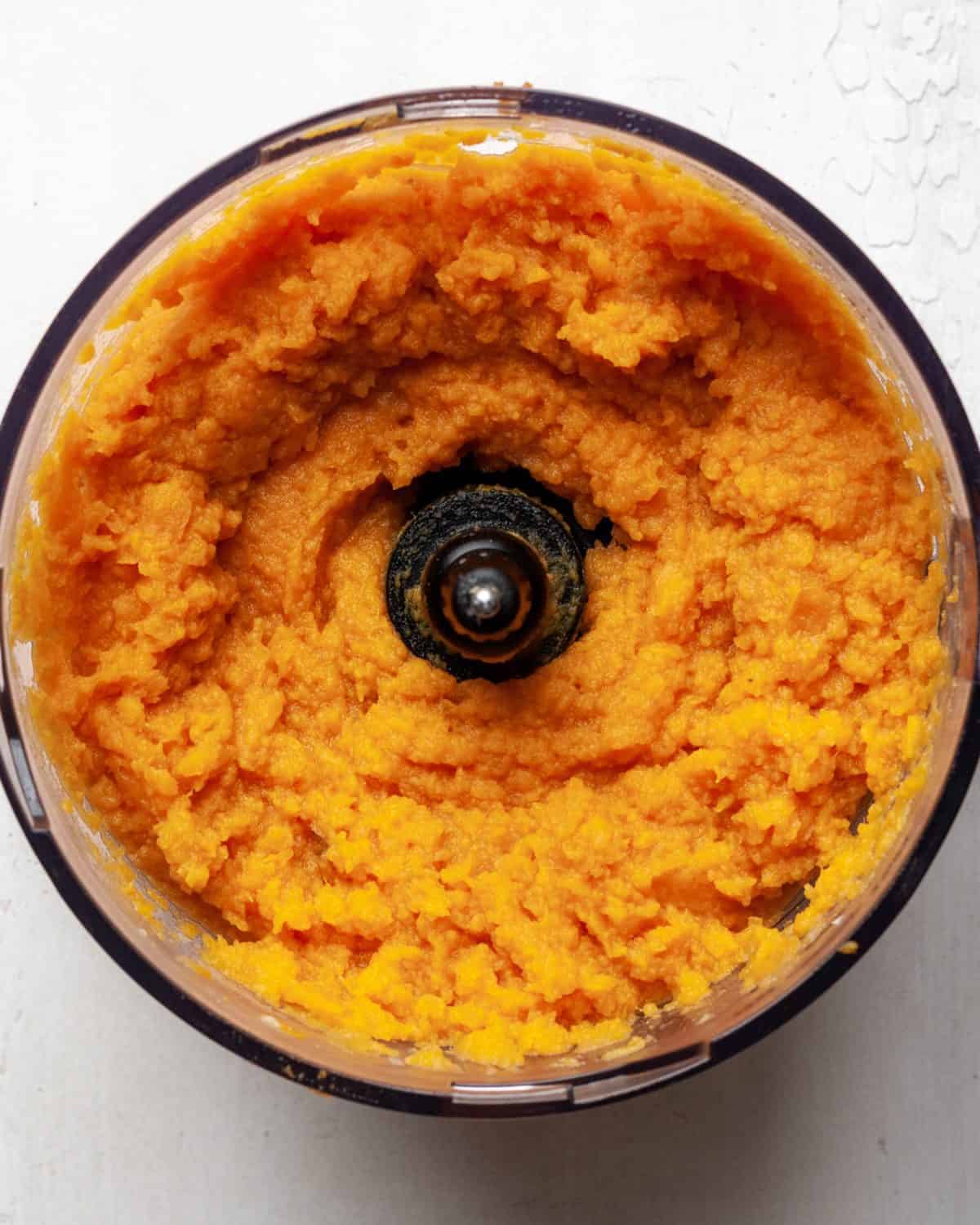 making healthy sweet potato casserole recipe - processed sweet potato puree in a food processor fitted with the "S" blade on a white surface.