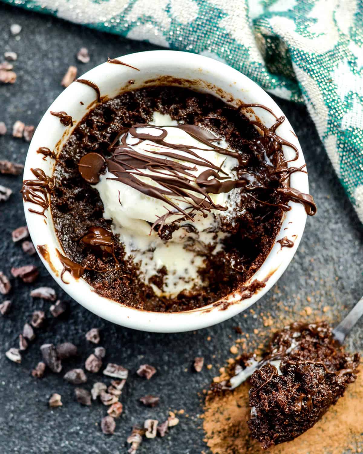 Overhead view of a healthy mug Brownie with paleo vanilla ice cream and a chocolate drizzle. A spoon with a bite of brownie on it is resting next to the ramekin. 