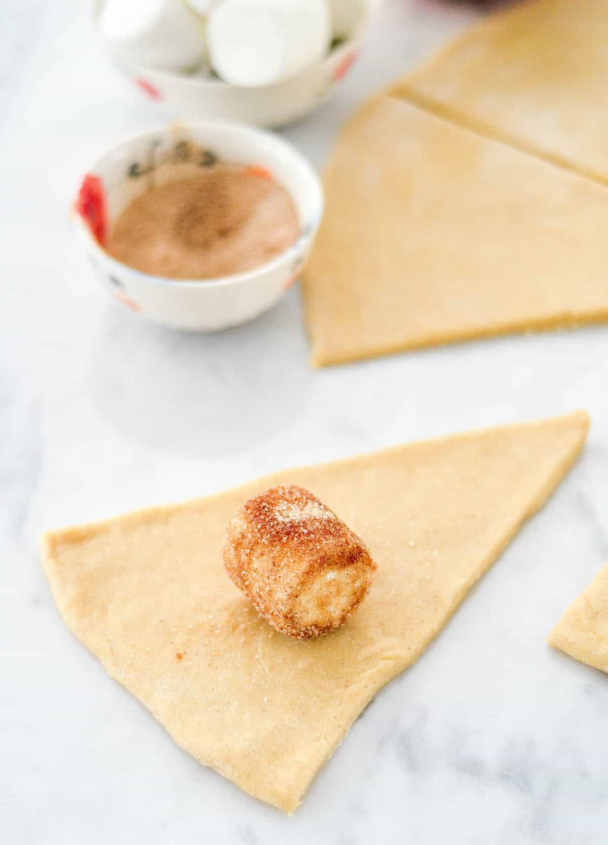 Side process shot of the homemade resurrection rolls showing a marshmallow that was dipped in butter and coated with cinnamon sugar on a triangular piece of dough ready to be rolled. 