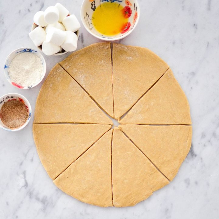 Overhead process shot of the homemade resurrection rolls dough rolled into a circle & cut into 8 triangular pieces with the other ingredients and a rolling pin situated around it. 