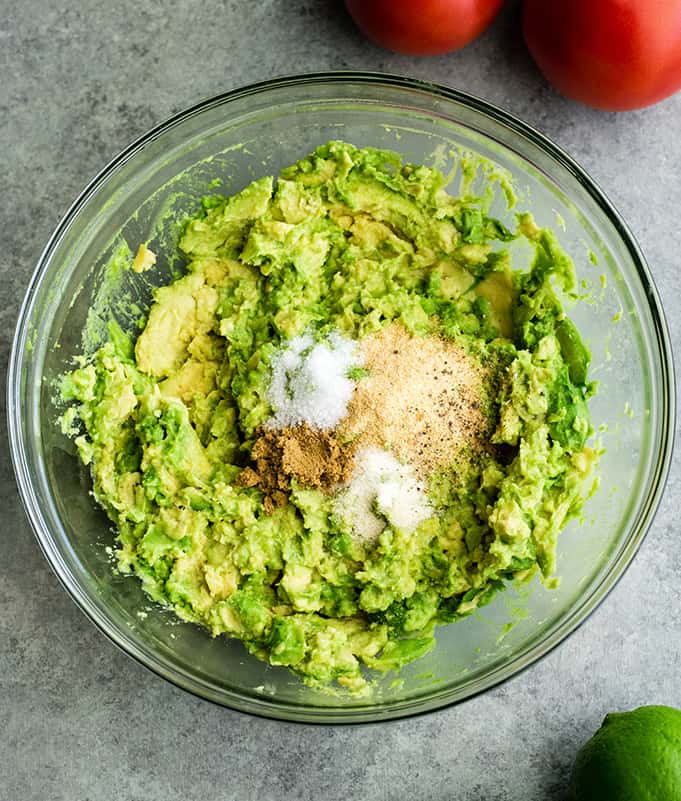 This quick & easy guacamole recipe is made with a handful of healthy ingredients and is ready in 5 minutes! This simple guacamole dip is our favorite appetizer and we always make it on taco nights! Watch the video to learn how to make this guacamole recipe! #guacamole #guacamolerecipe #tacos #appetizer #dip overhead view of the spices being added to mashed avocado in a glass bowl to make easy guacamole recipe
