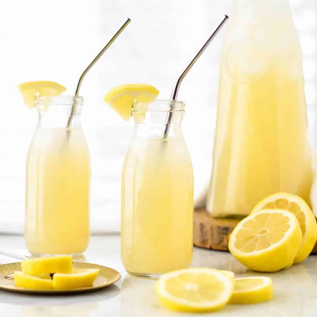 Front view of two glass bottles filled with Healthy Lemonade with Orange Blossom water with the pitcher of healthy lemonade in the background
