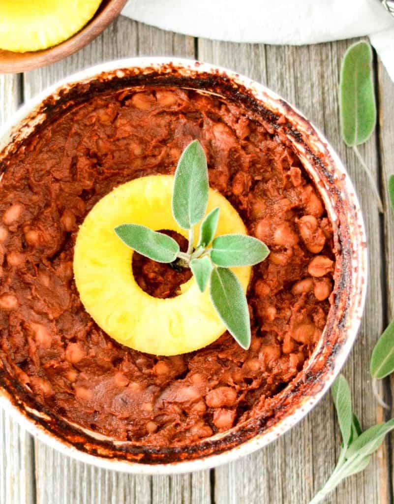 Overhead view of a round baking dish of Healthy Vegan Baked Beans garnished with a pineapple ring and fresh sage