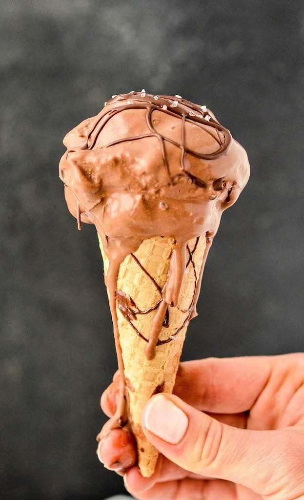 Front view of a hand holding a sugar cone with paleo chocolate ice cream dripping down the cone drizzled with chocolate topped with sea salt