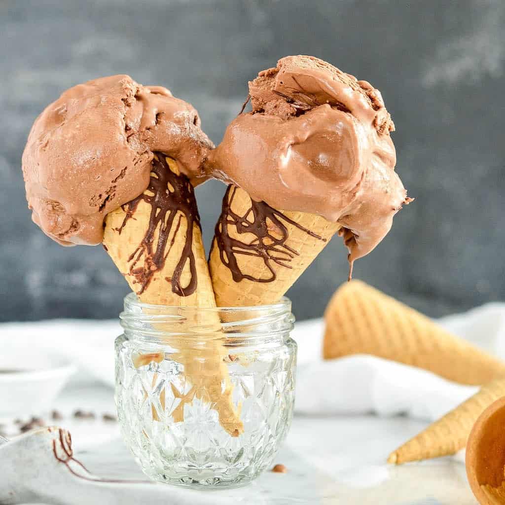 front view of two sugar cones drizzled with chocolate with scoops of paleo chocolate ice cream on them with the ice cream dripping down 
