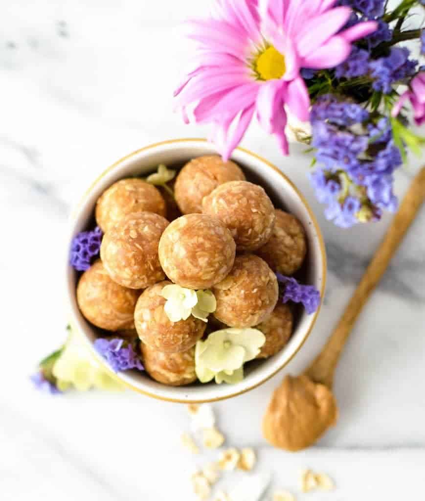 Overhead view of Peanut Butter Oatmeal Balls in a small white & gold bowl with purple flowers in a vase nearby
