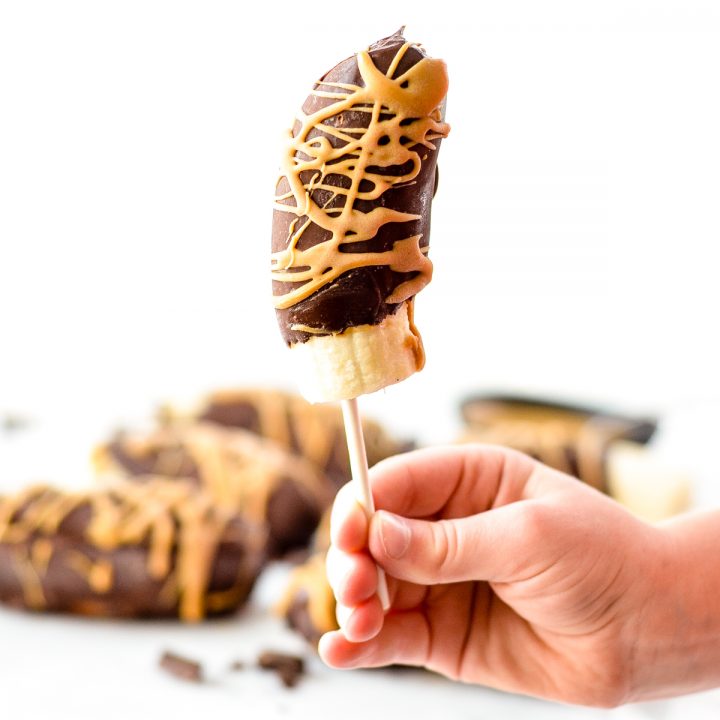 Front view of a hand holding one Chocolate Covered frozen Bananas on a stick with more in the background