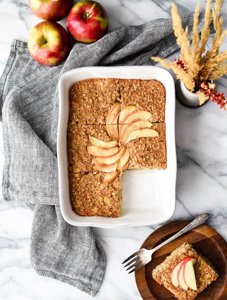 Overhead view of a rectangular baking dish with Apple Cinnamon Baked Oatmeal cut into 6 pieces, one piece is removed on a plate next to the pan
