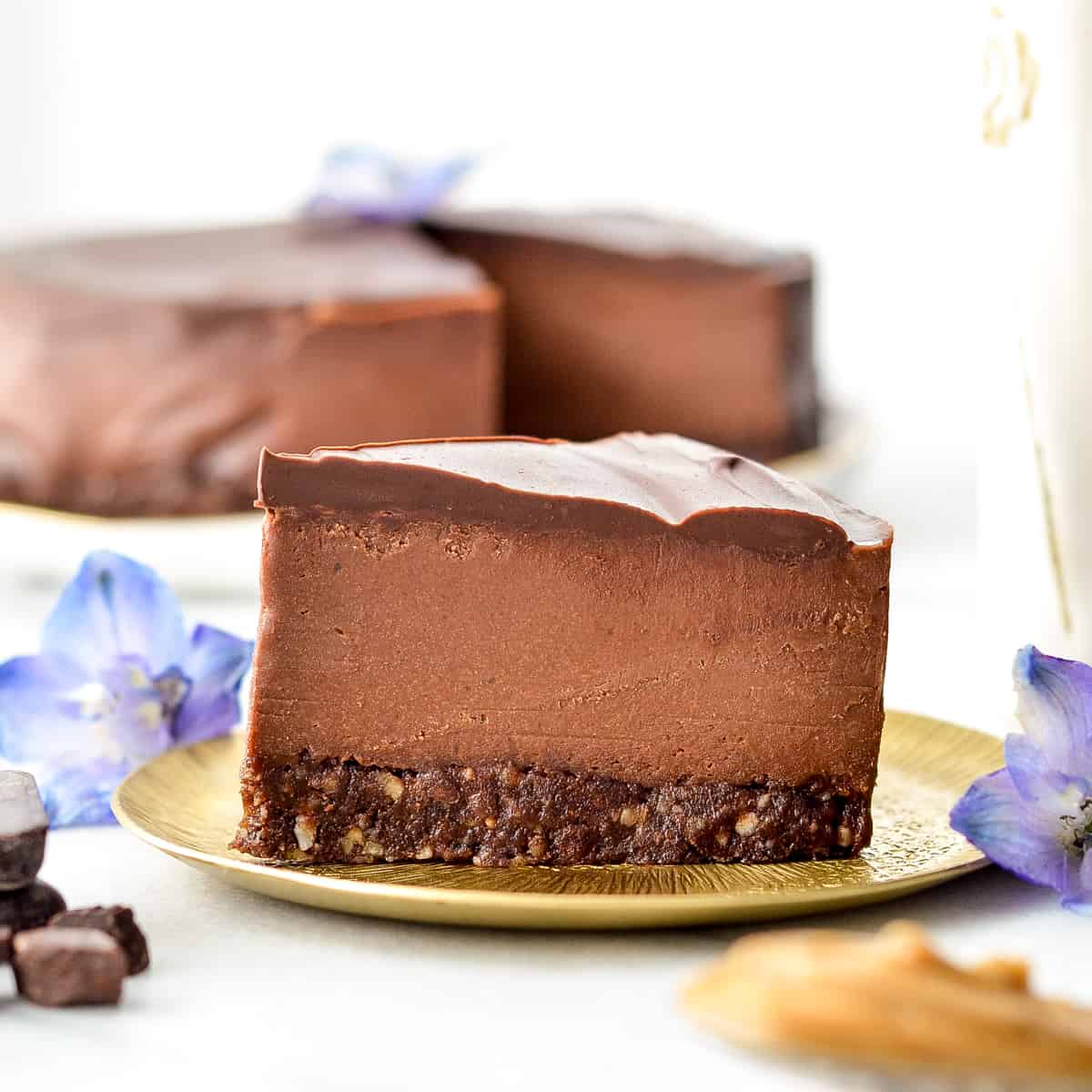 Side view of a slice of No-Bake Vegan Chocolate Peanut Butter Cheesecake on a plate with the rest of the cheesecake in the background