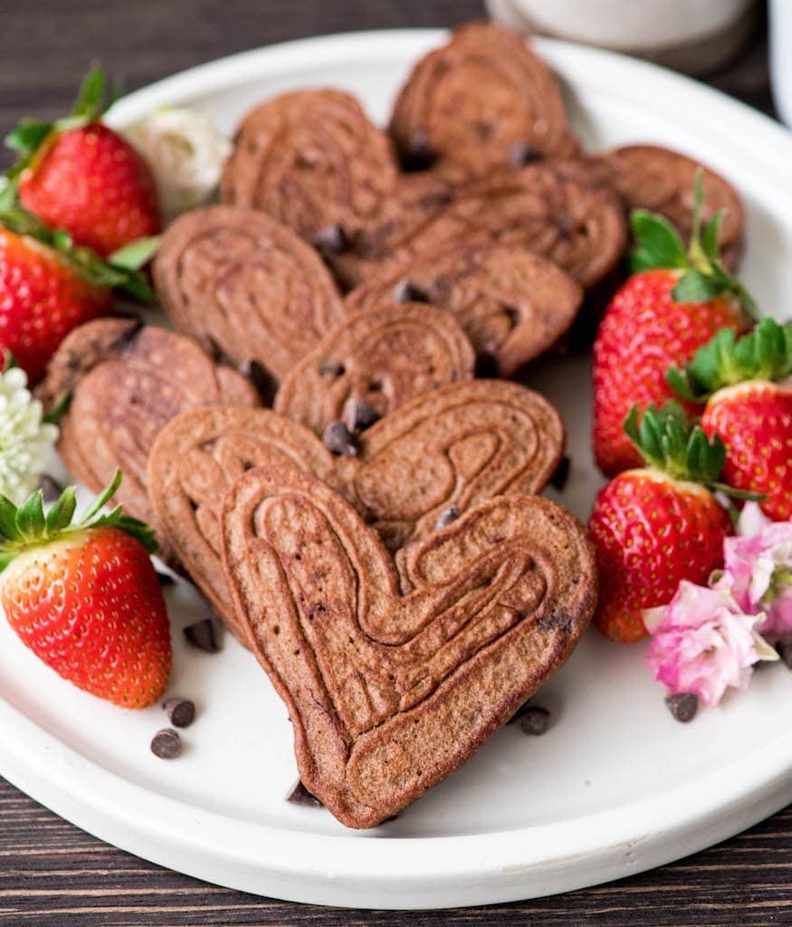 Overhead view of a 6 heart-shaped Gluten-Free Healthy Chocolate Pancakes Recipe on a white circular plate surrounded by strawberries, flowers and chocolate chips 