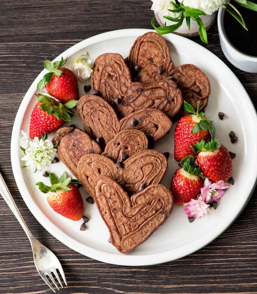 Overhead view of a 6 heart-shaped Healthy Chocolate Pancakes Recipe on a white circular plate surrounded by strawberries, flowers and chocolate chips 
