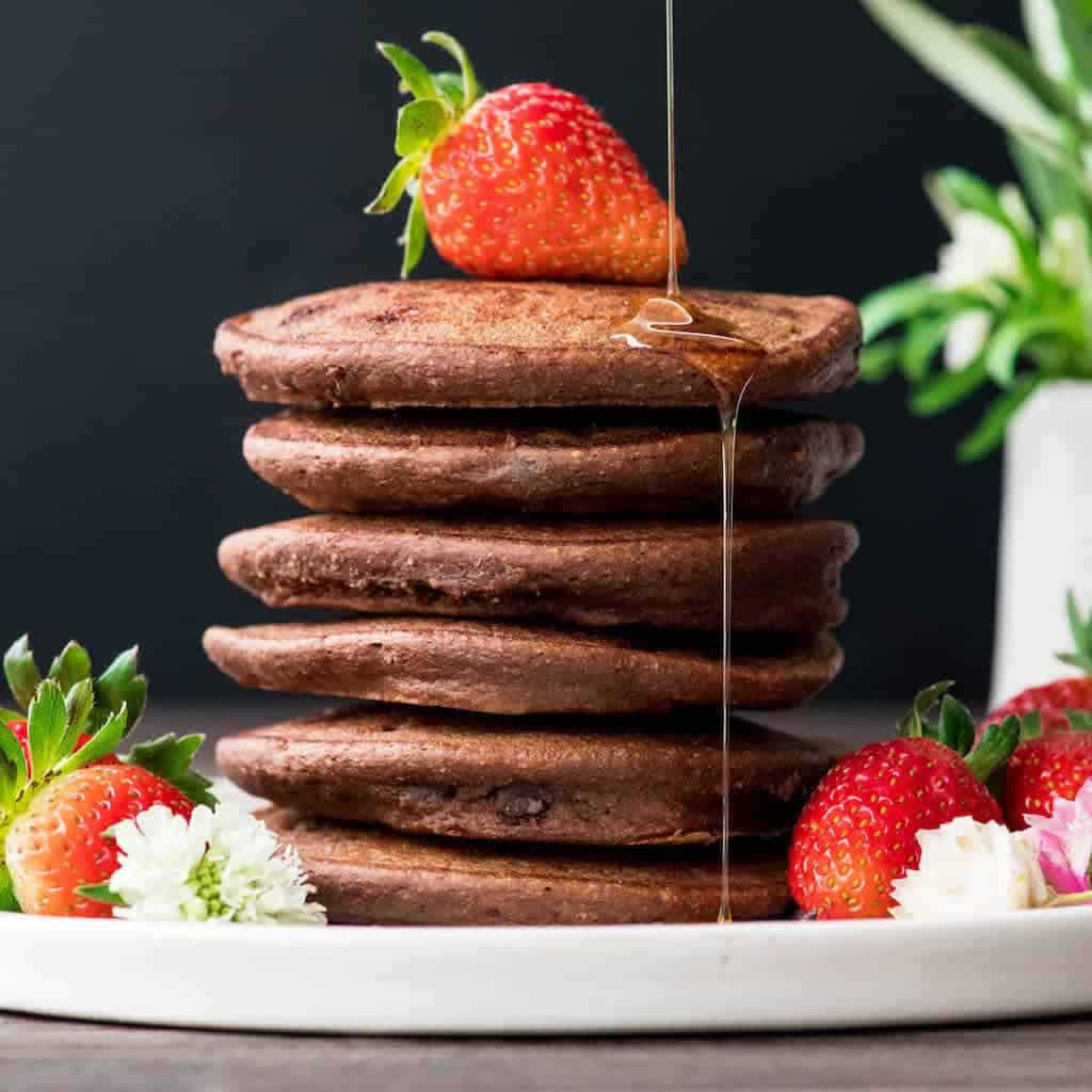 Front view of a stack of 6 Gluten-Free Healthy Chocolate Pancakes Recipe with a strawberry on top and maple being poured onto the stack and dripping down in one place