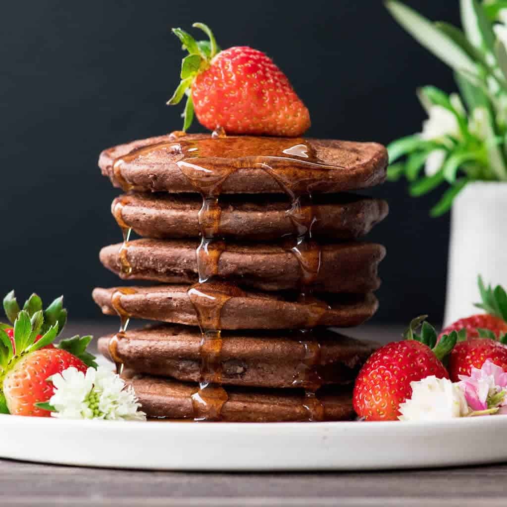 Front view of a stack of 6 Gluten-Free Healthy Chocolate Pancakes Recipe with a strawberry on top and maple syrup dripping down the sides in three places