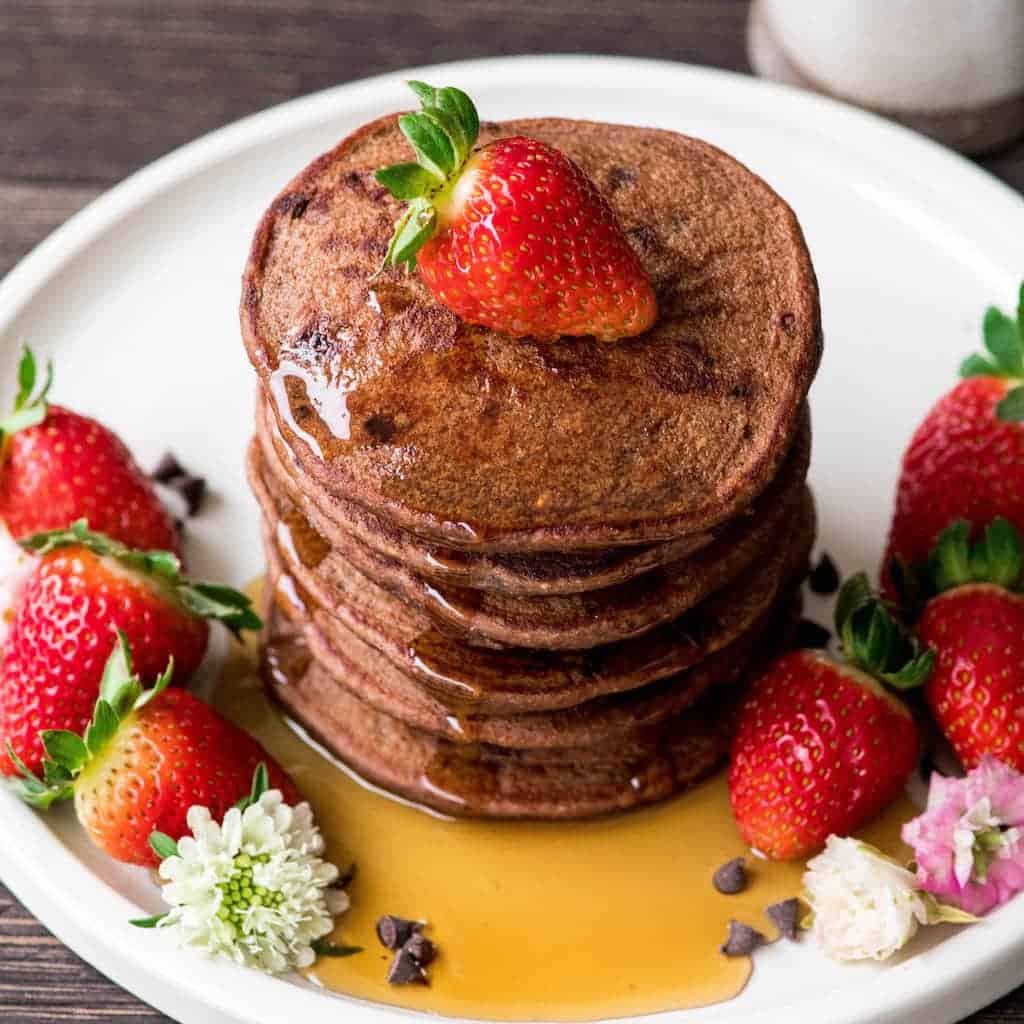 Overhead side view of a stack of 6 Gluten-Free Healthy Chocolate Pancakes Recipe with a strawberry on top and maple syrup dripping down the sides in three places
