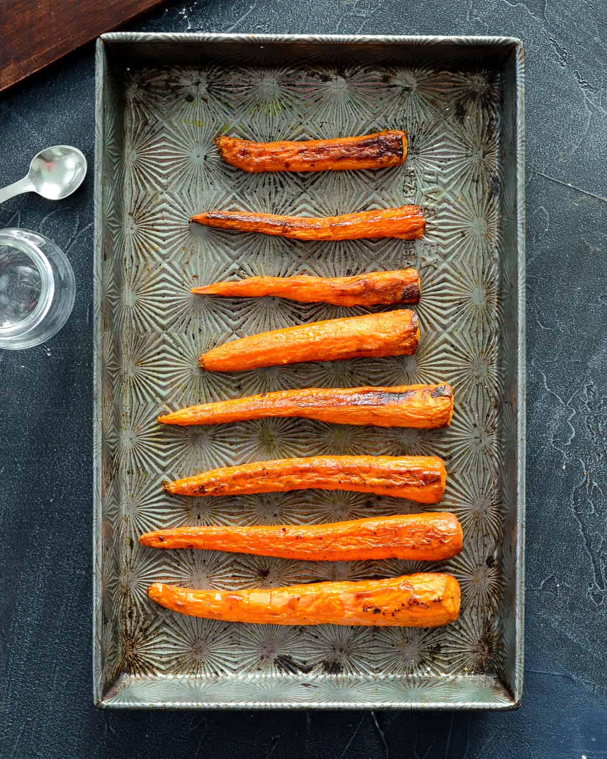 Overhead view of a pan of roasted carrots with a glass baby food jar and spoon next to it in the making of homemade baby food carrots