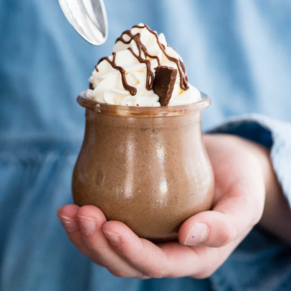 Front view of a hand holding a jar of Creamy Chocolate Peanut Butter Chia Pudding while the other hand is holding a spoon above it ready to dig in 