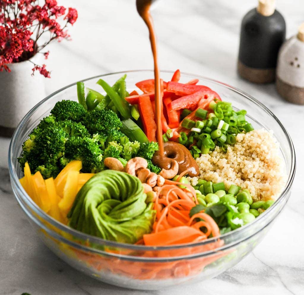 zijaanzicht van pindasaus wordt gegoten in een kom met alle ingrediënten van Aziatische quinoa salade met pinda dressing Recept