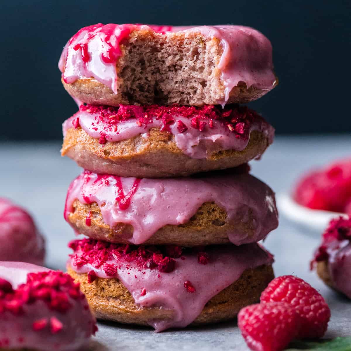 Front view of a stack of four raspberry baked donuts with other donuts laying around them. The top donut has a bite taken out of it. 