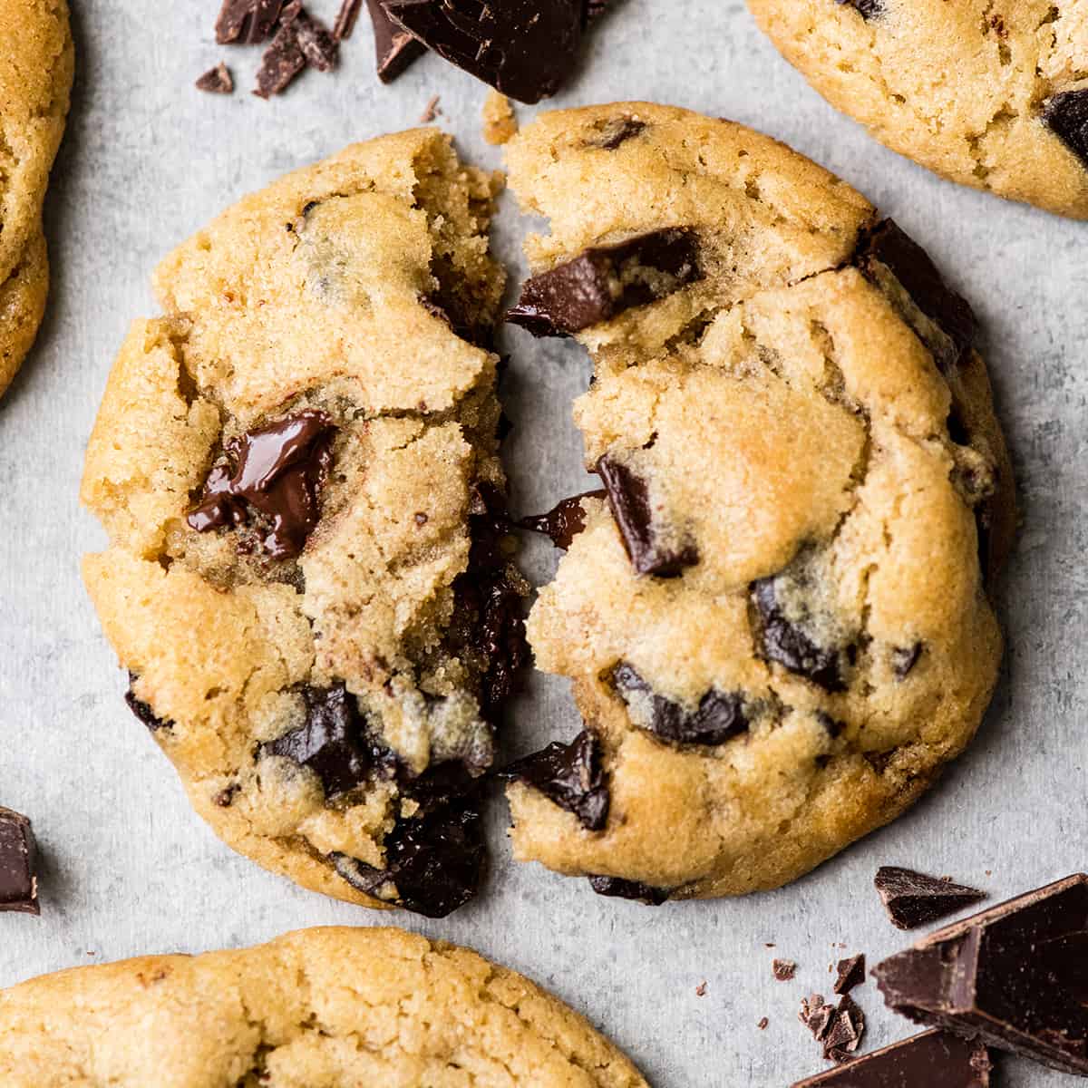 Overhead view of one chocolate chip cookie torn in half with melty chocolate chips surrounded by three other cookies and chopped chocolate