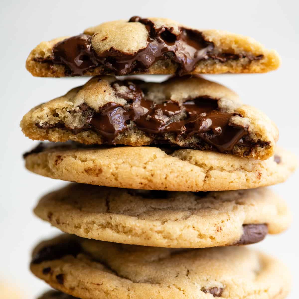 a stack of 4 chocolate chip cookies, the top one is cut in half and both halves are stacked on each other so they gooey texture and melty chocolate chips are visible.