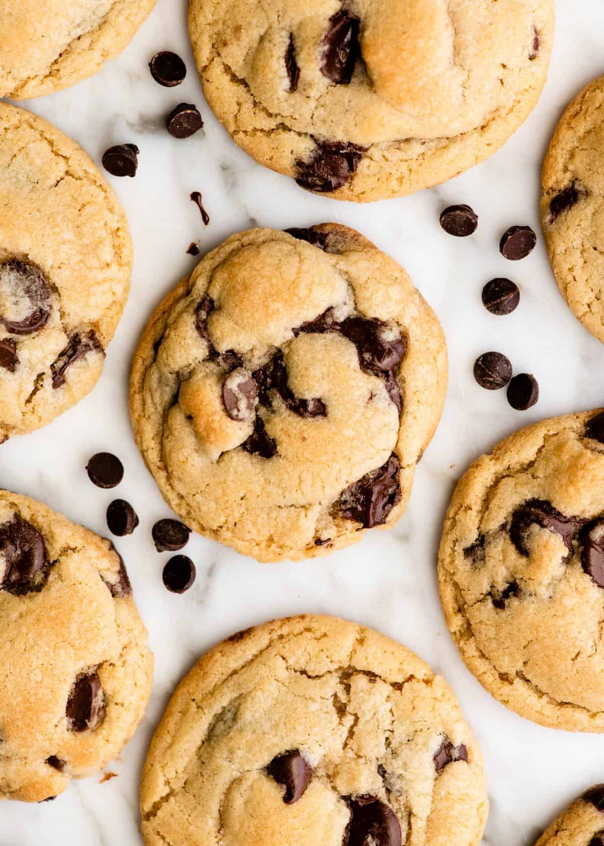 7 chocolate chip cookies on a marble surface surrounded by chocolate chips.