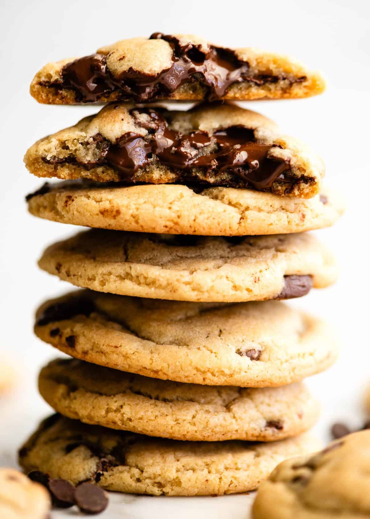 a stack of 6 chocolate chip cookies, the top one is cut in half and both halves are stacked on each other so they gooey texture and melty chocolate chips are visible.