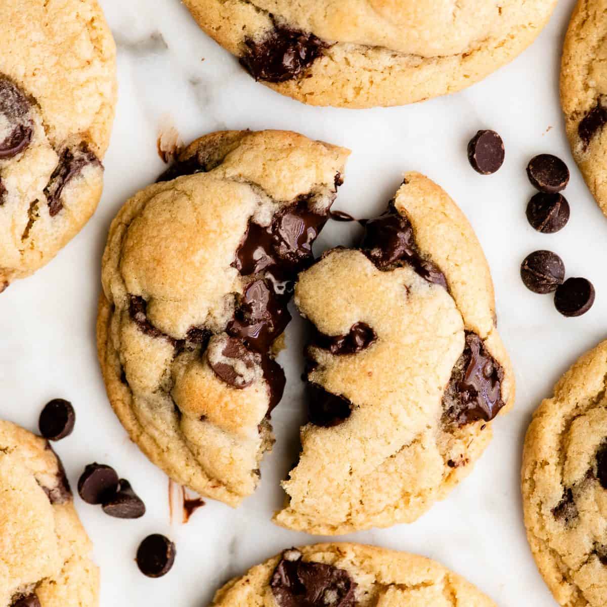 one chocolate chip cookie torn in half so the melty chocolate chips are visible, surrounded by 6 other best chocolate chip cookies and piles of chocolate chips all on a marble surface