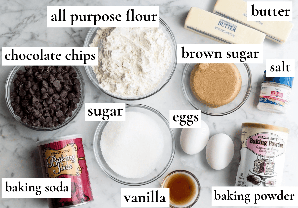 overhead photo of the ingredients in this chocolate chip cookie recipe - all labeled some in bowls on a marble surface. The ingredients are butter, brown sugar, salt, baking powder eggs, vanilla, sugar, baking soda, chocolate chips and flour