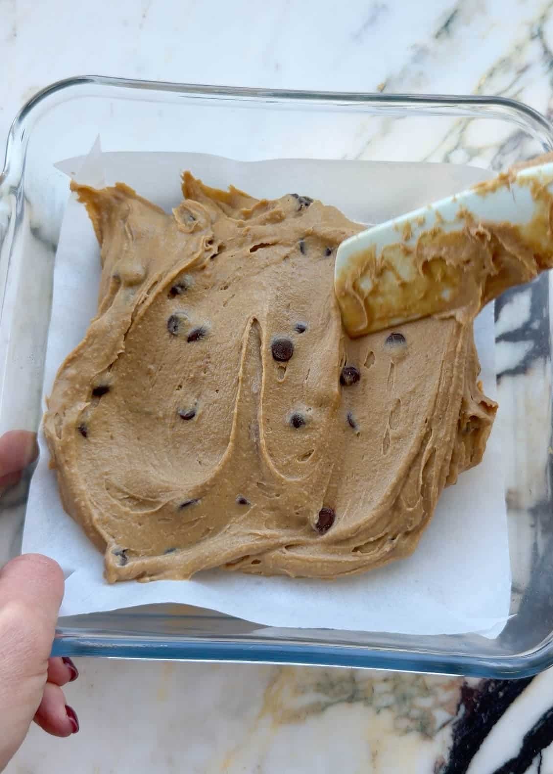 chickpea blondies batter being spread into a square glass baking dish lined with parchment paper.