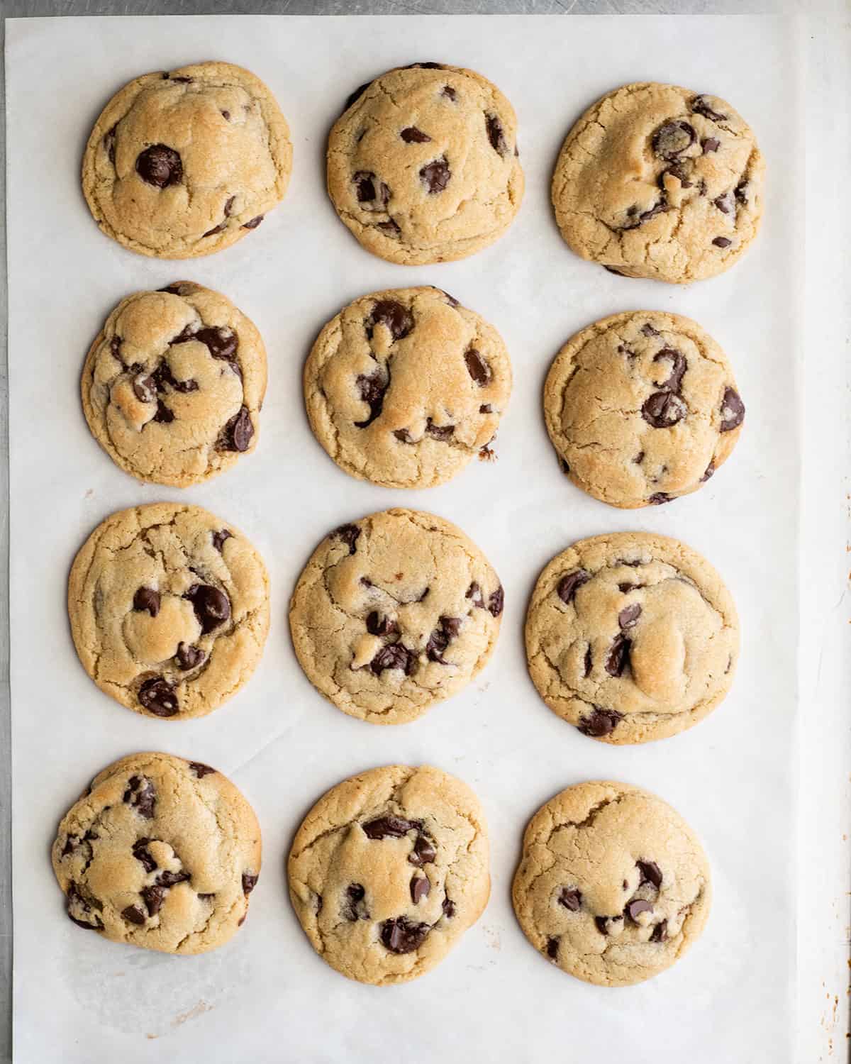 how to make chocolate chip cookies - 12 cookie chocolate chip cookies arranged in 4 rows of 3 on a baking sheet lined with white parchment paper after baking.