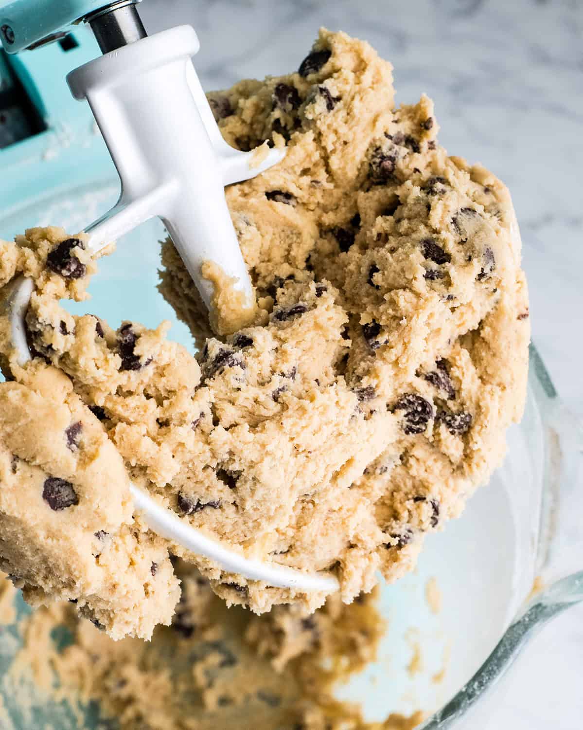 how to make chocolate chip cookies - the finished dough on the white paddle attachment in a glass mixing bowl of a light blue kitchen aid mixer making the best chocolate chip cookies recipe
