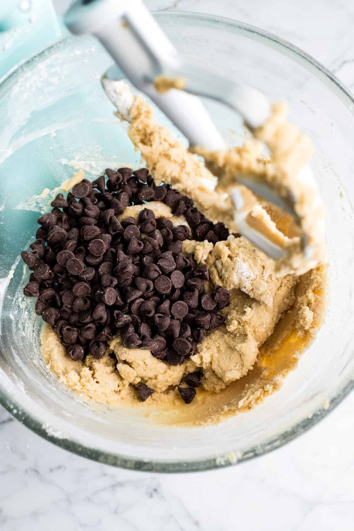 how to make chocolate chip cookies - chocolate chips added to the dough in a glass mixing bowl of a light blue kitchen aid mixer fitted with a white paddle attachment before mixing together making the best chocolate chip cookies recipe
