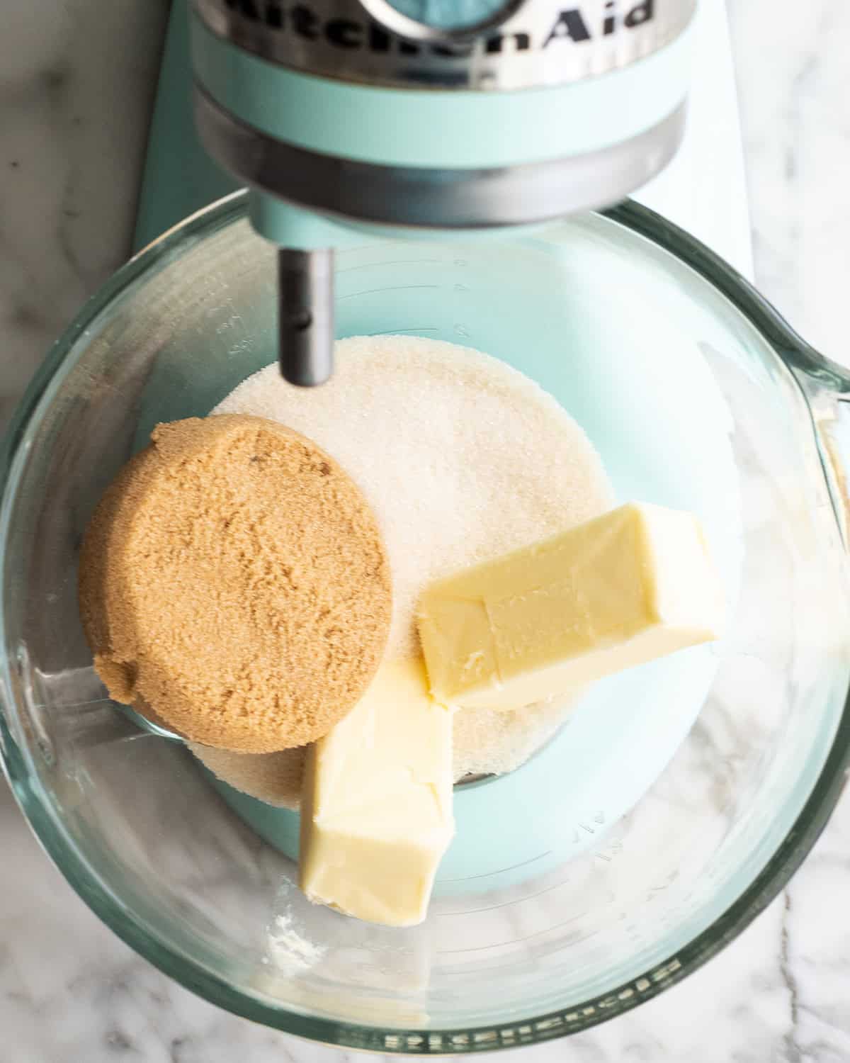 how to make chocolate chip cookies - two sticks of butter, white sugar and brown sugar in a glass mixing bowl of a light blue kitchen aid mixer before beating together making the best chocolate chip cookies recipe