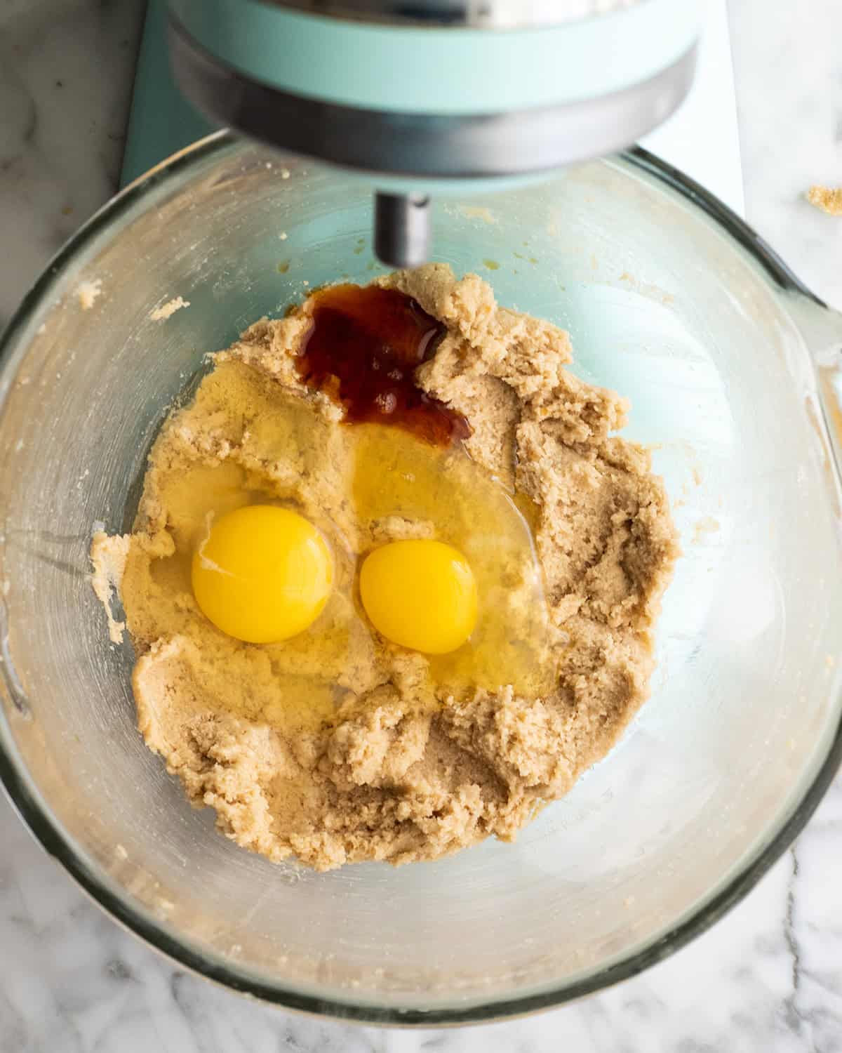 how to make chocolate chip cookies - beaten butter and sugar mixture with two eggs and a splash of vanilla added in a glass mixing bowl of a light blue kitchen aid mixer before beating together making the best chocolate chip cookies recipe