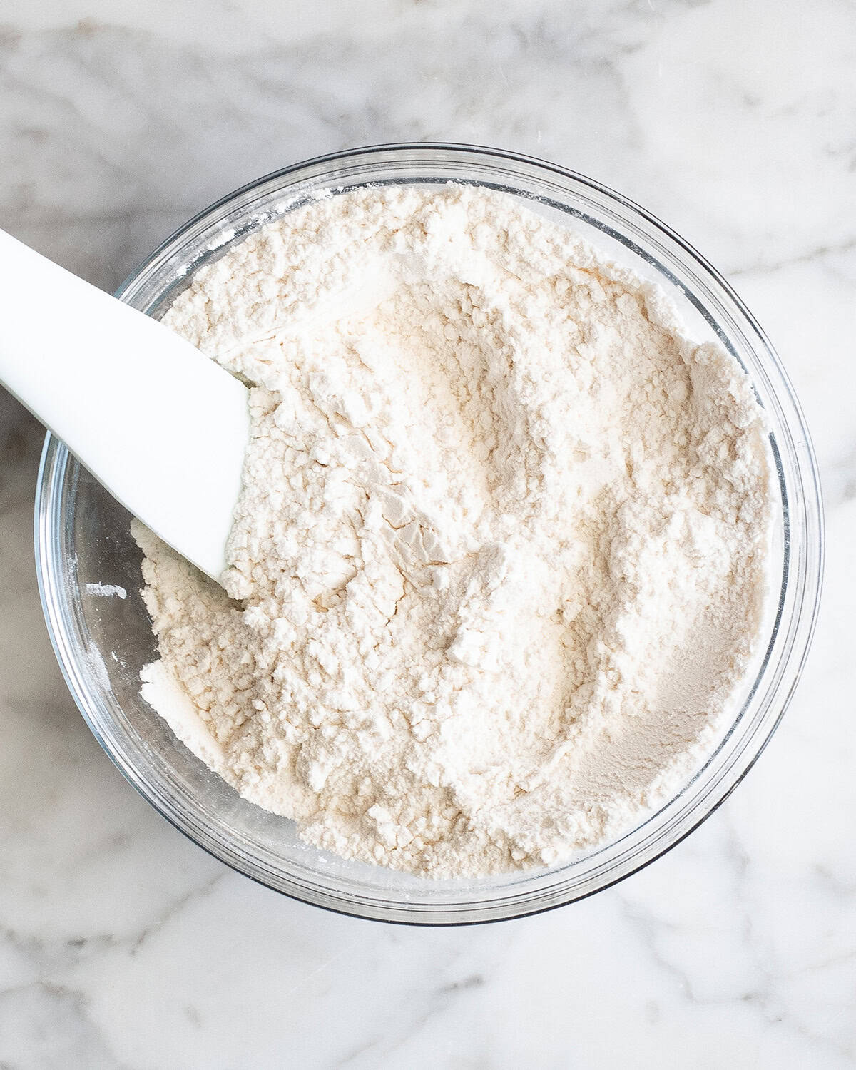 how to make chocolate chip cookies - flour, baking soda, baking powder and salt in a glass bowl with a very light blue silicone spatula after being stirred together on a marble surface making the best chocolate chip cookies recipe