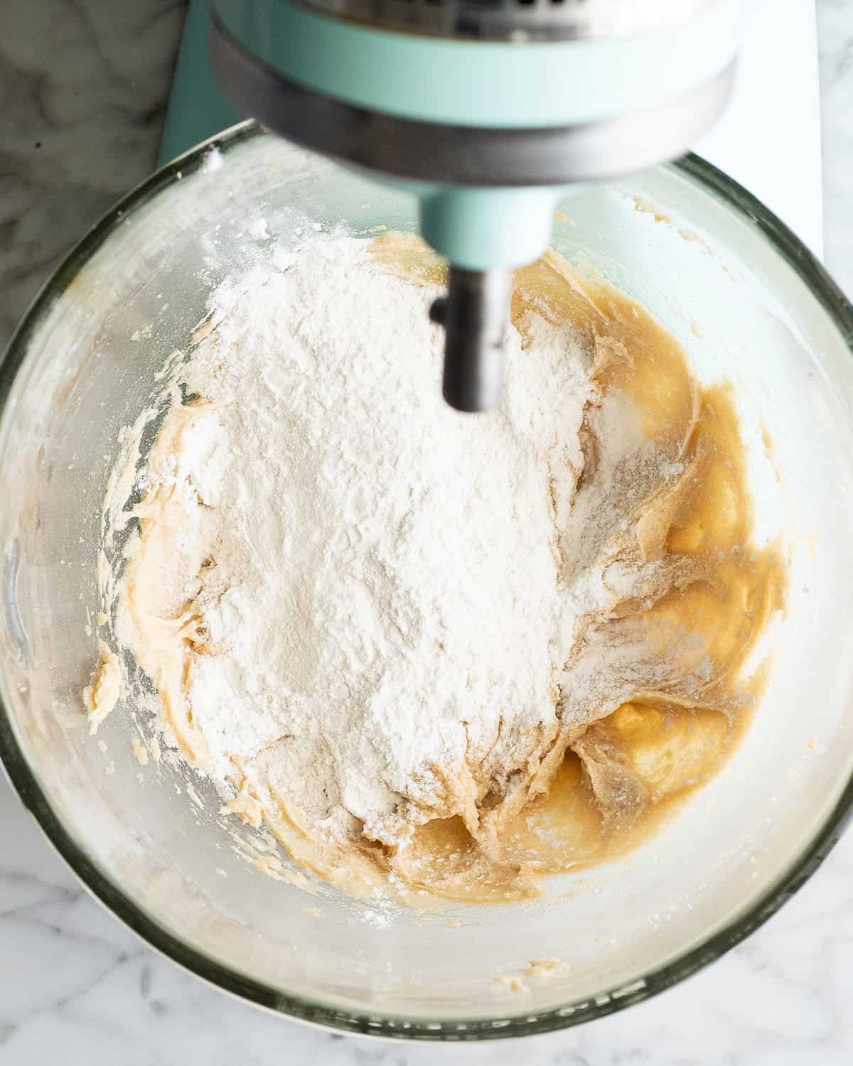 how to make chocolate chip cookies -flour mixture added to the beaten wet ingredients in a glass mixing bowl of a light blue kitchen aid mixer before beating together making the best chocolate chip cookies recipe