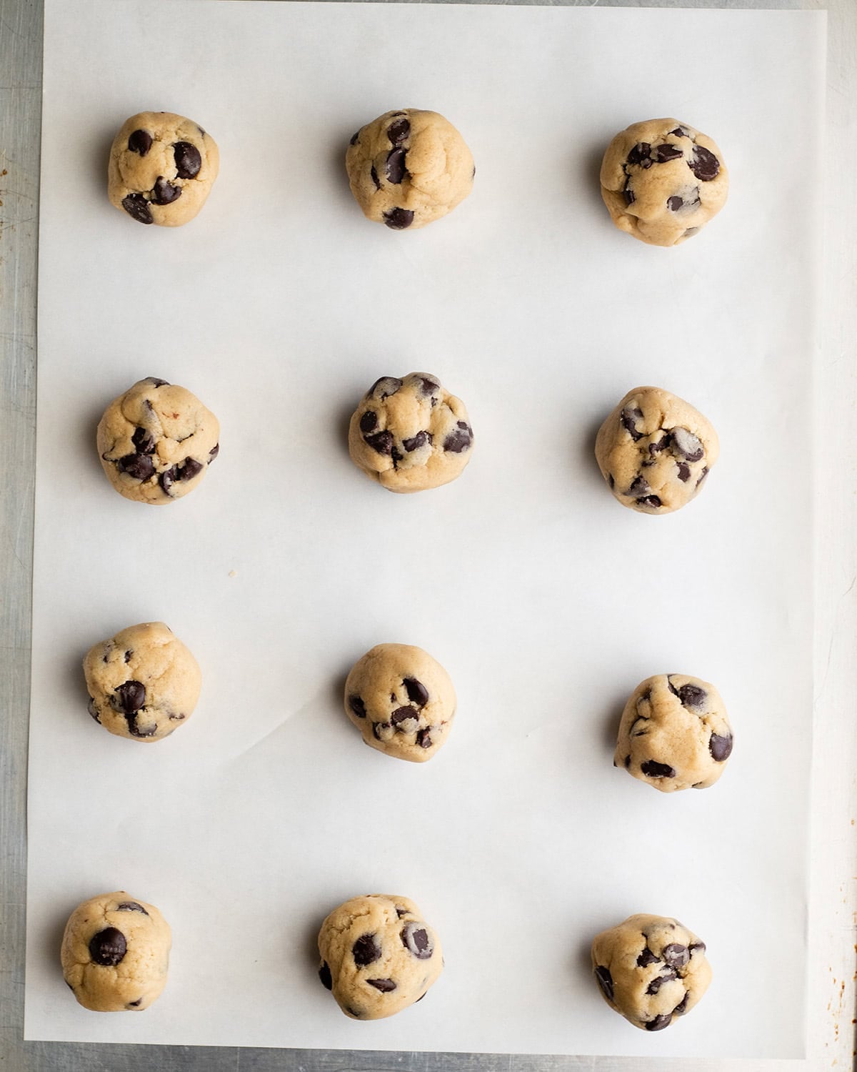 how to make chocolate chip cookies - 12 cookie dough balls arranged in 4 rows of 3 on a baking sheet lined with white parchment paper before baking