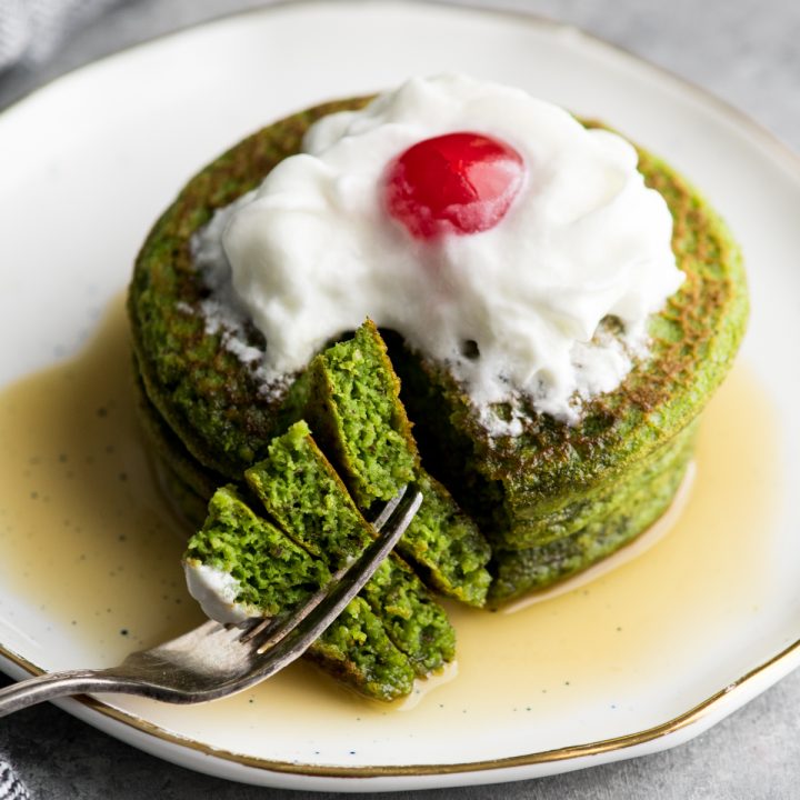 a stack of three spinach pancakes topped with whipped cream and a cherry in a pool of syrup with one bite cut out and on a fork. 