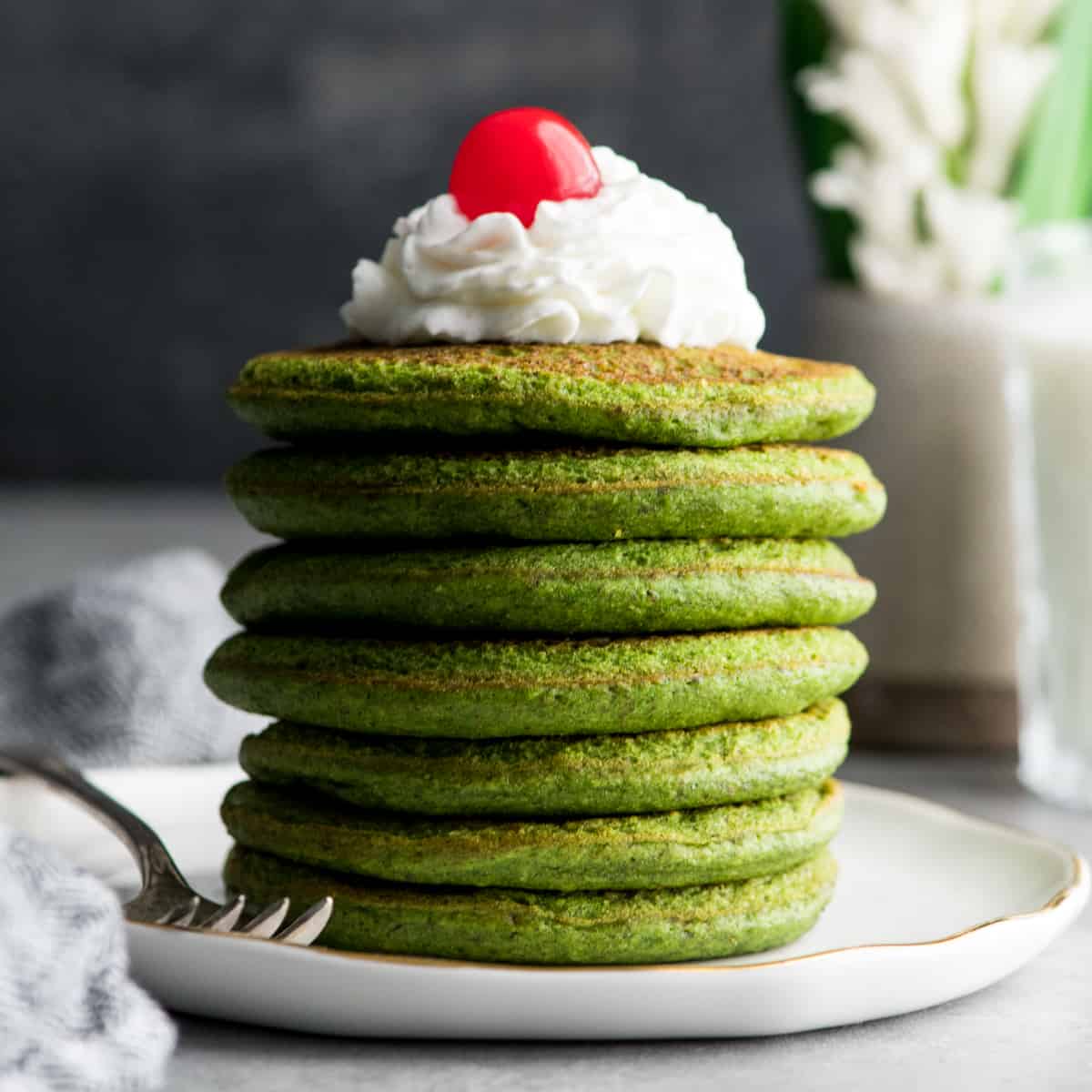 front view of a stack fo 7 healthy spinach pancakes topped with whipped cream & a cherry 