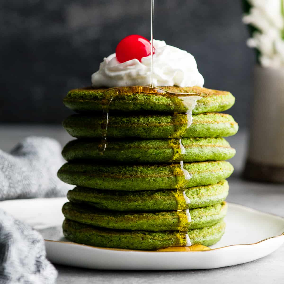 front view of a stack fo 7 healthy spinach pancakes topped with whipped cream & a cherry being drizzled with maple syrup