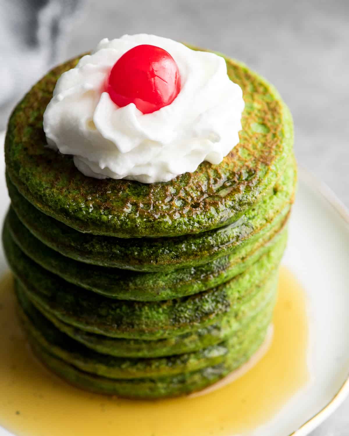 Up-close overhead/angled view of a stack of 7 spinach pancakes topped with whipped cream and a cherry sitting in a pool of syrup