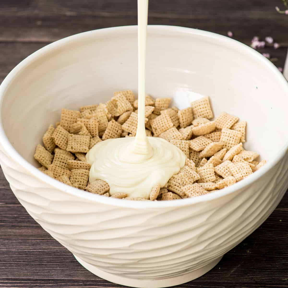 Side view of white chocolate being poured into a mixing bowl filled with rice chex making White Chocolate Matcha Puppy Chow 